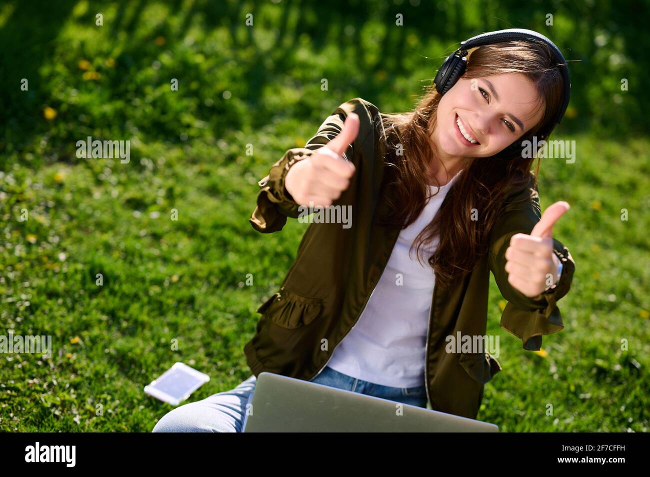 Sorridente studente donna attraente seduta sull'erba all'aperto utilizzando laptop e cuffie e mostra il movimento del pollice verso l'alto Foto Stock
