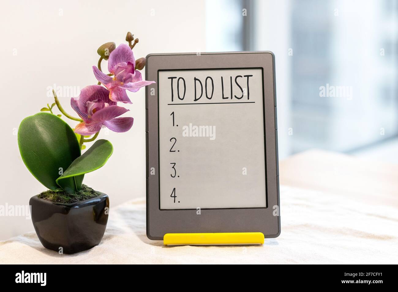 Elenco delle attività in un tablet grigio su un supporto giallo accanto ad un'orchidea rosa artificiale sul tavolo da cucina con una tovaglia di lino vicino alla finestra Foto Stock
