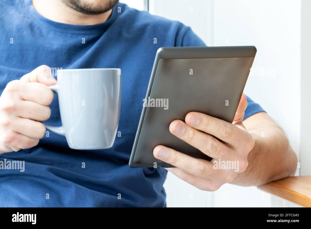 Un uomo in una t-shirt blu tiene un libro elettronico grigio e-reader in una mano e una tazza nell'altra. Leggere un libro o notizie su un tablet a colazione. SEL Foto Stock