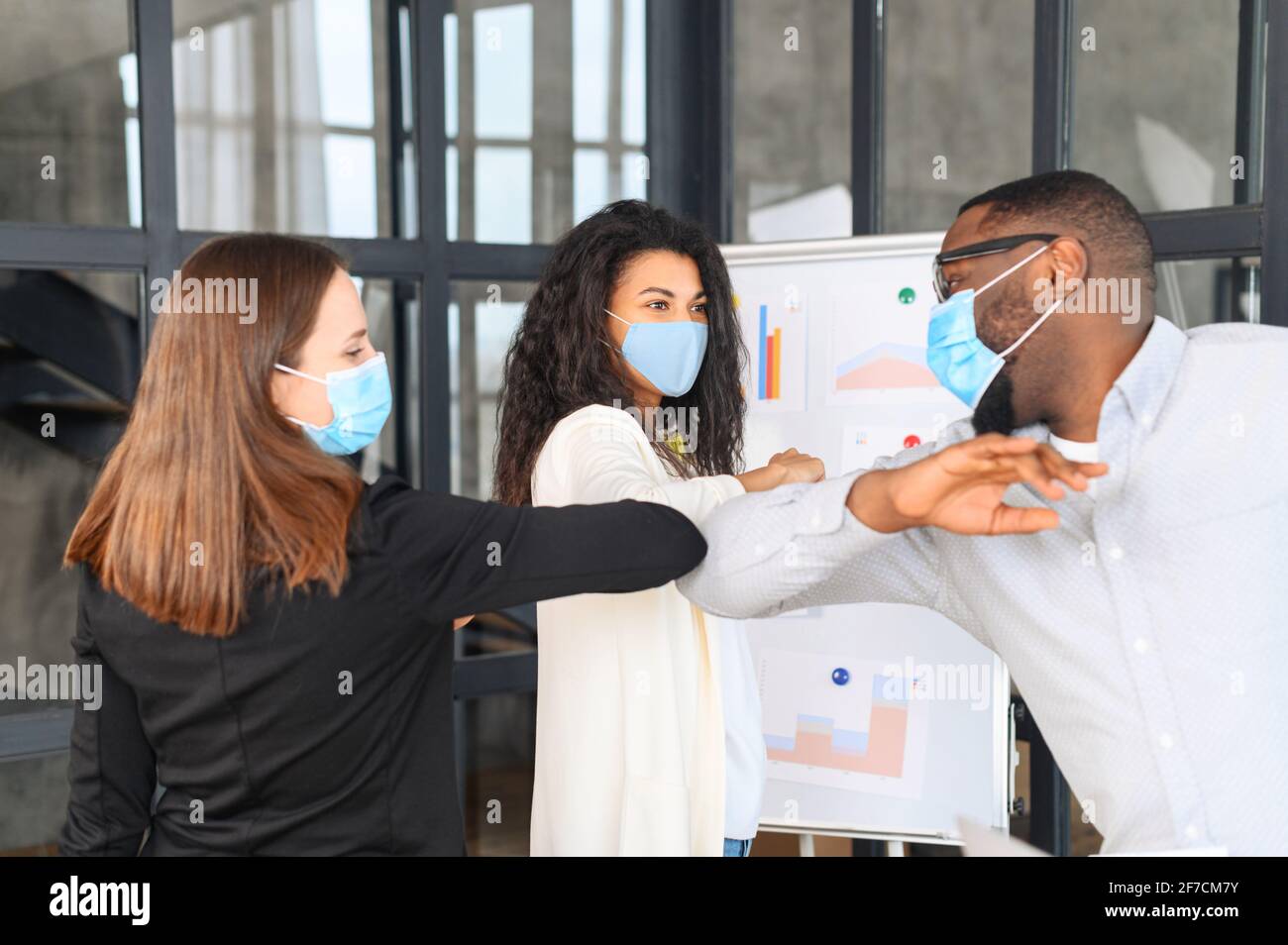 Diverse due donne e l'uomo, colleghi in maschere facciali di protezione saluto urtare gomiti sul posto di lavoro, donna collaboratori in coperture facciali proteggere da COVID-19 coronavirus in ufficio, concetto di assistenza sanitaria Foto Stock