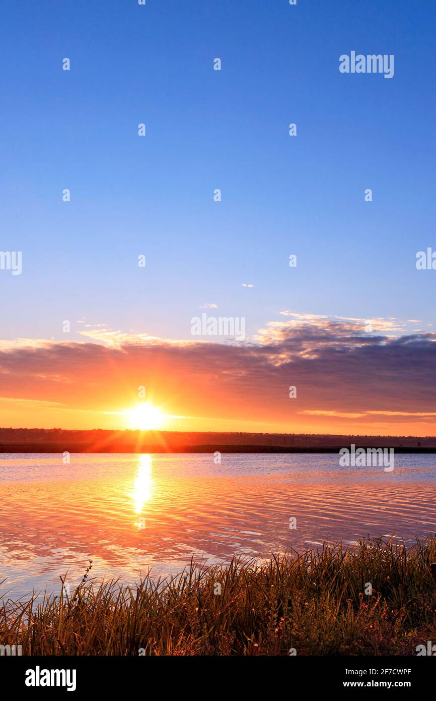 Alba mattutina sul fiume, il luminoso sole che sorge sopra l'orizzonte, i raggi del sole si riflettono nella superficie ondulata dell'acqua. Foto Stock