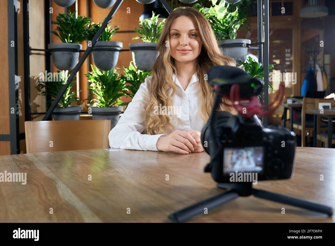 Ritratto di giovane donna sorridente con bei capelli lunghi seduti al tavolo e dando un'intervista in un moderno cafè. Concetto di comunicazione di processo tramite telecamera. Foto Stock