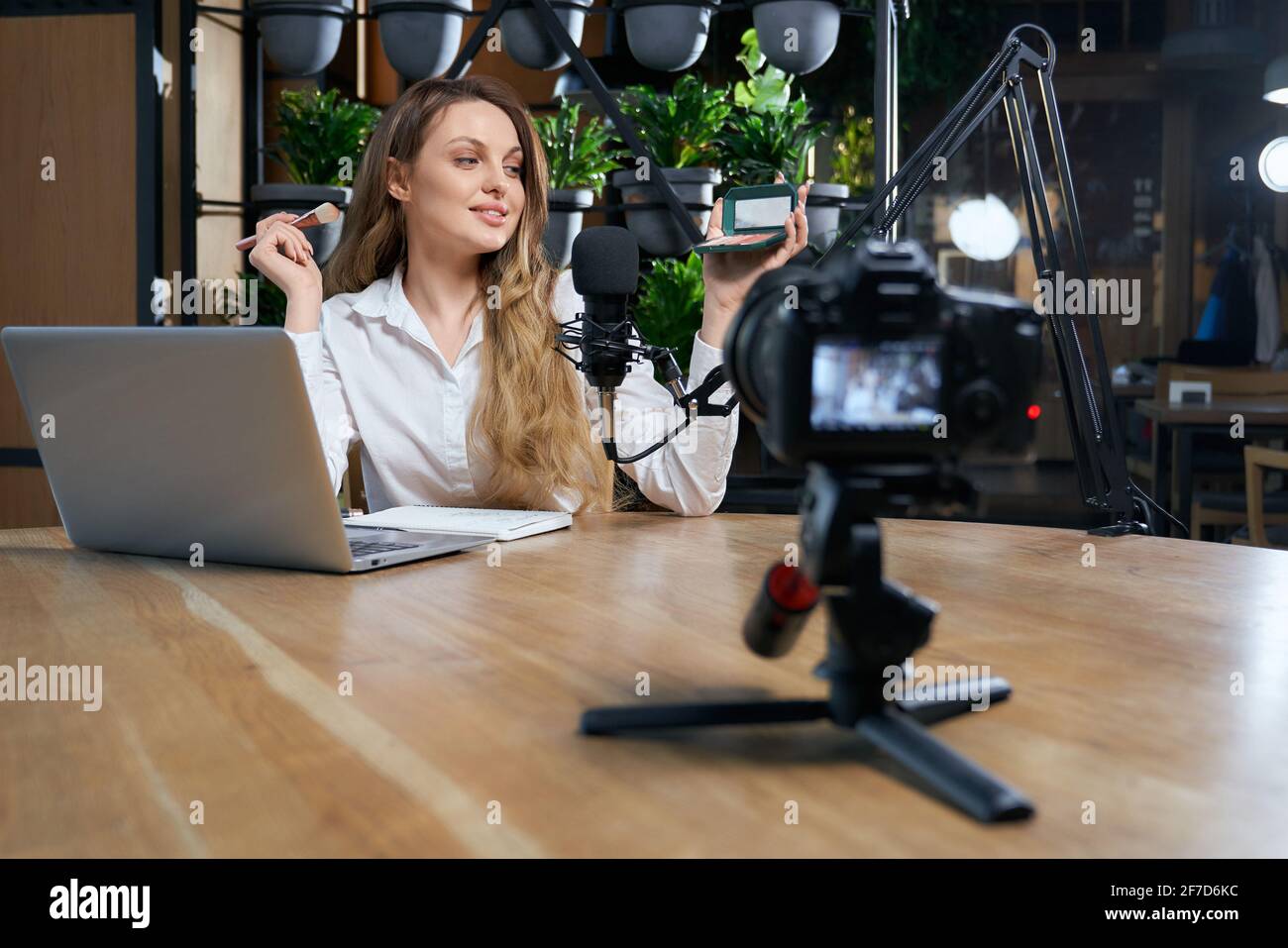 Vista frontale di una giovane bella donna seduta al tavolo vicino al computer portatile e al microfono nel bar. Concetto di blogger preparazione per l'intervista e fare il trucco con buon umore. Foto Stock