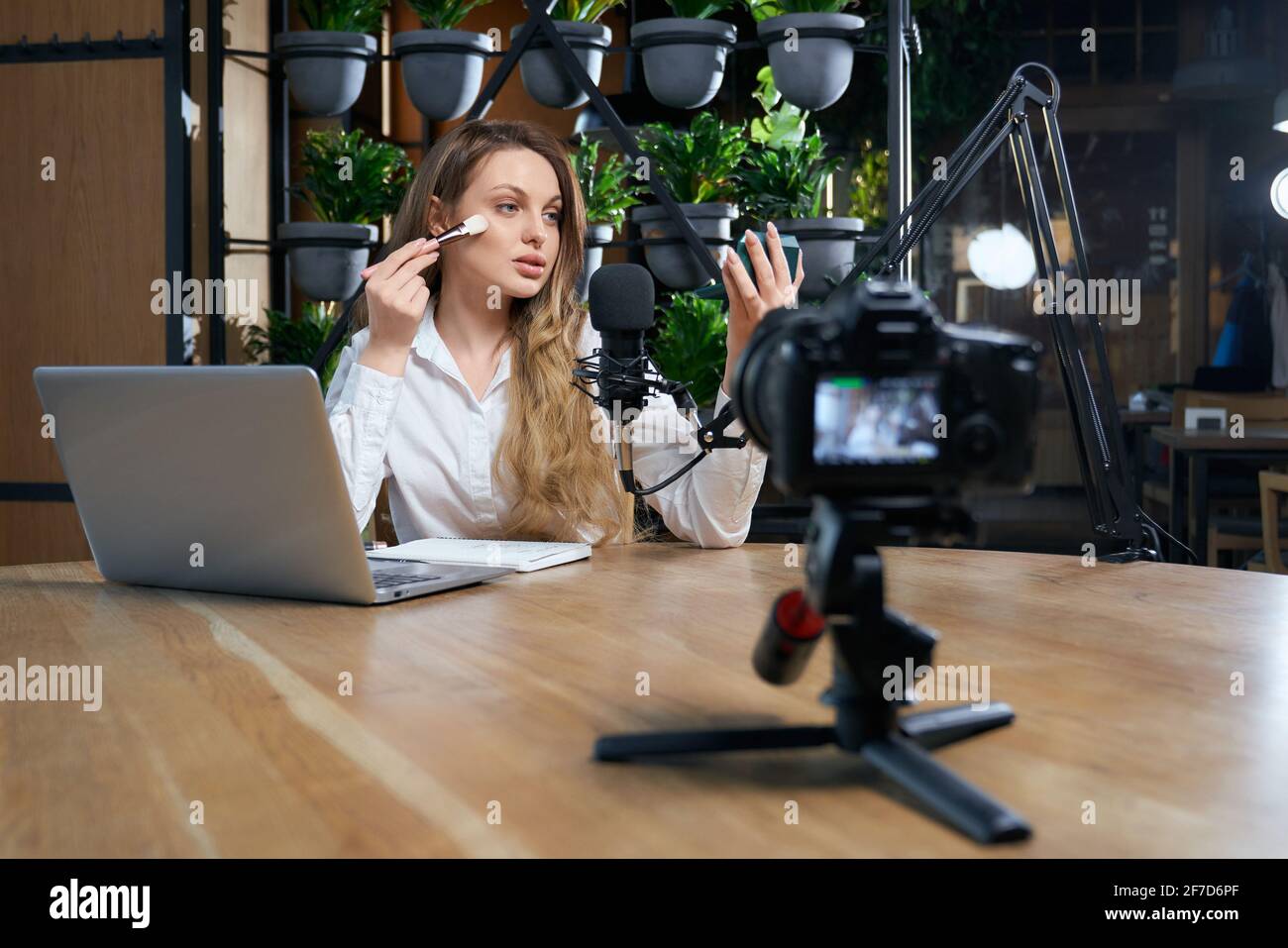 Vista frontale della giovane bella donna con i capelli lunghi guardando in piccolo specchio e facendo il trucco al tavolo nel caffè. Concetto di processo preparazione per l'intervista con laptop e microfono. Foto Stock