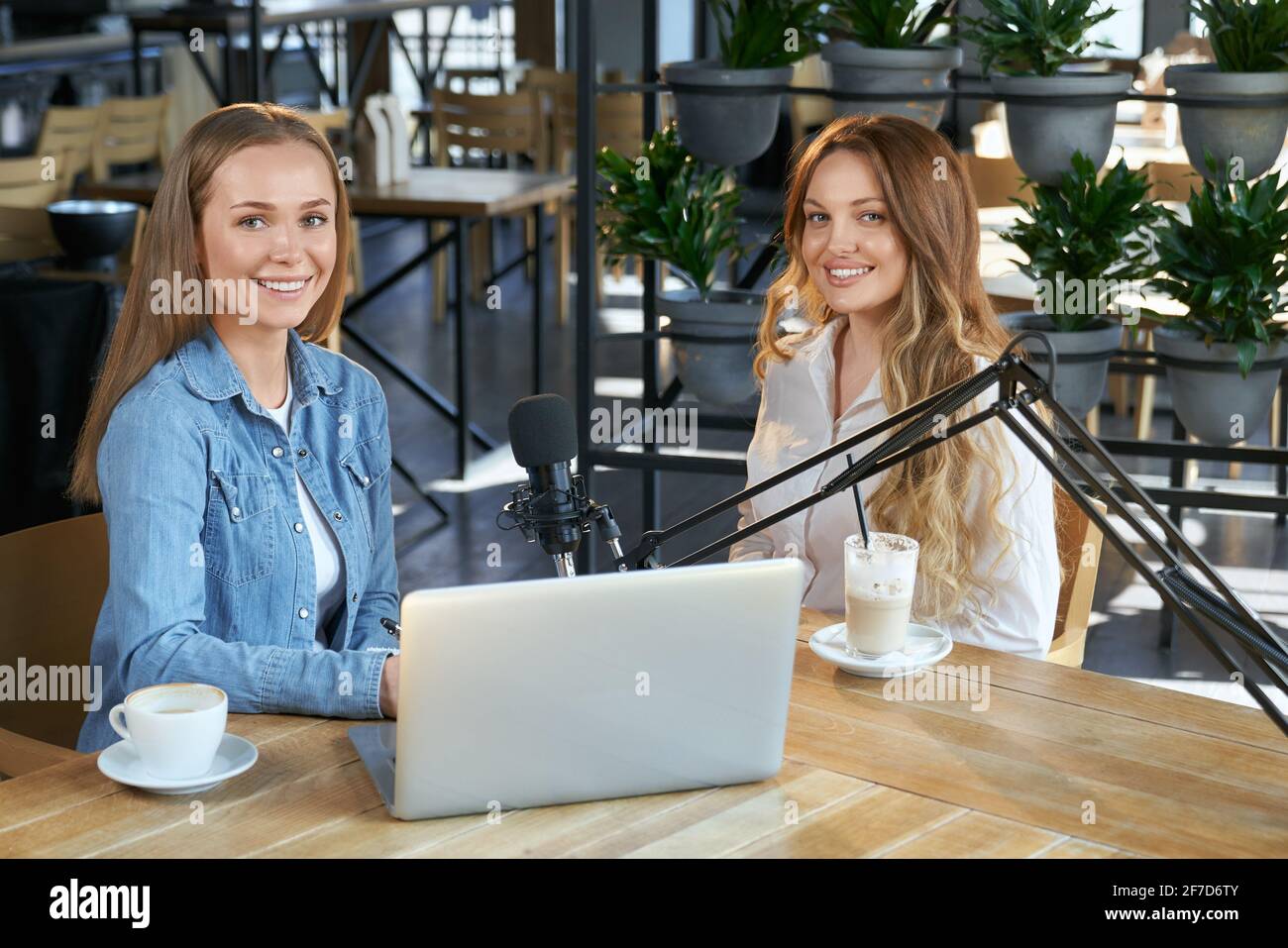 Vista frontale di due belle giovani blogger donne seduti in caffetteria con gustoso caffè e parlare con i seguaci online. Concetto di comunicazione dei blogger con gli abbonati tramite trasmissione su laptop. Foto Stock