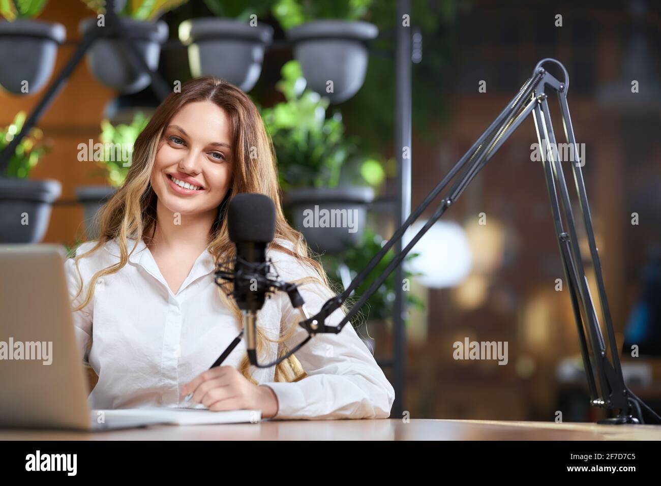 Primo piano ritratto di sorridente giovane bella blogger donna in camicia bianca registrare nuovo video con microfono per seguaci. Concetto di processo di comunicazione con gli abbonati online. Foto Stock