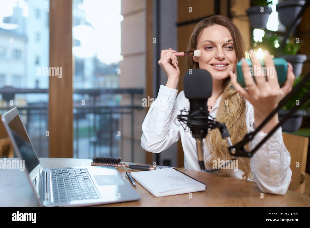 Primo piano ritratto di sorridente giovane attraente donna con capelli lunghi in camicia bianca facendo trucco e guardando specchio. Concetto di processo di preparazione per la trasmissione live con i follower. Foto Stock