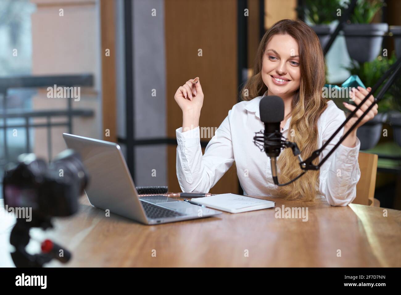 Ritratto di sorridente giovane blogger donna in camicia bianca che parla con i seguaci nel microfono dal laptop. Concetto di comunicazione con i follower in trasmissione live. Foto Stock