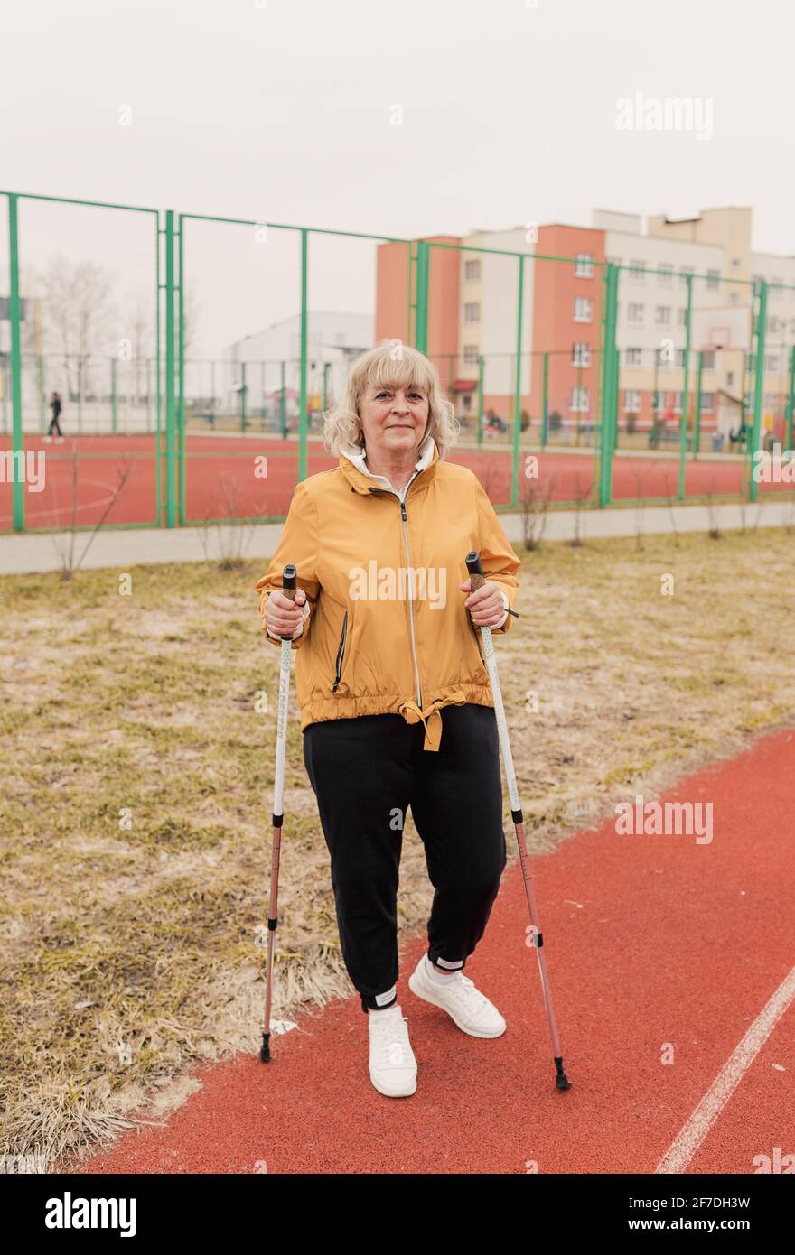 Una donna anziana in una giacca gialla si alza con le levette nordiche in mano allo stadio su un tapis roulant rosso. Stile di vita sano Foto Stock