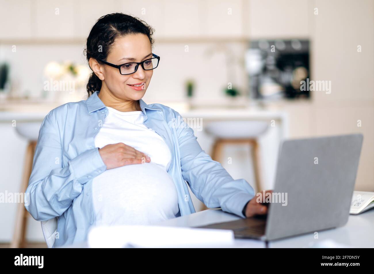 Concentrata gioiosa donna di razza mista incinta indossare occhiali, seduto al tavolo, utilizza il laptop per lavorare o navigare in rete, ottenere informazioni sul lavoro, delicatamente stracciando la pancia, sorridendo Foto Stock