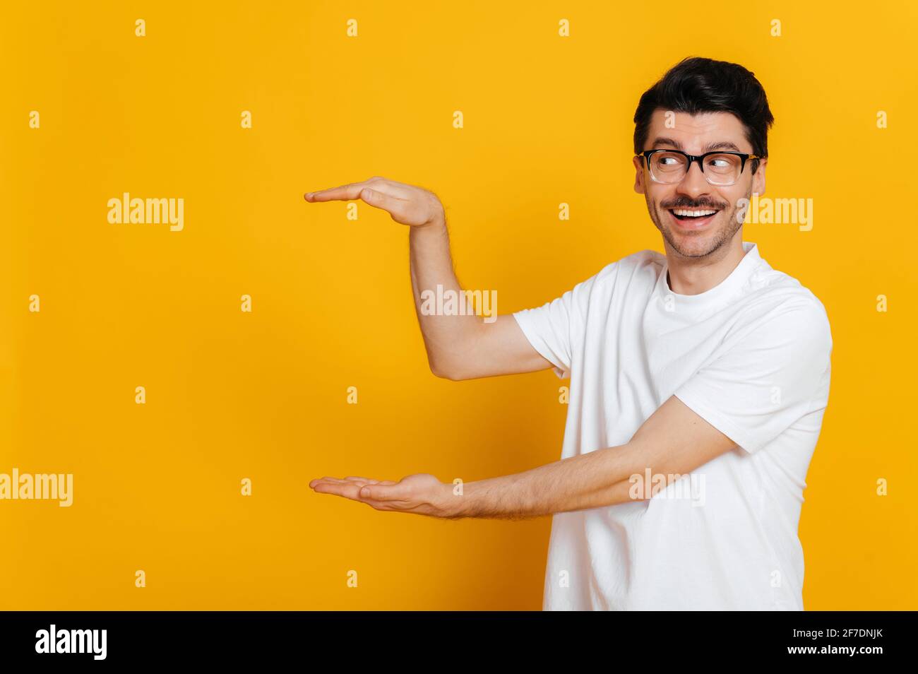 Eccitato stupito felice caucasico unshaven ragazzo in bianco t-shirt e occhiali guarda e mostrando con le mani a uno spazio vuoto per la tua presentazione, in piedi su isolato sfondo arancione, toothy sorridente Foto Stock