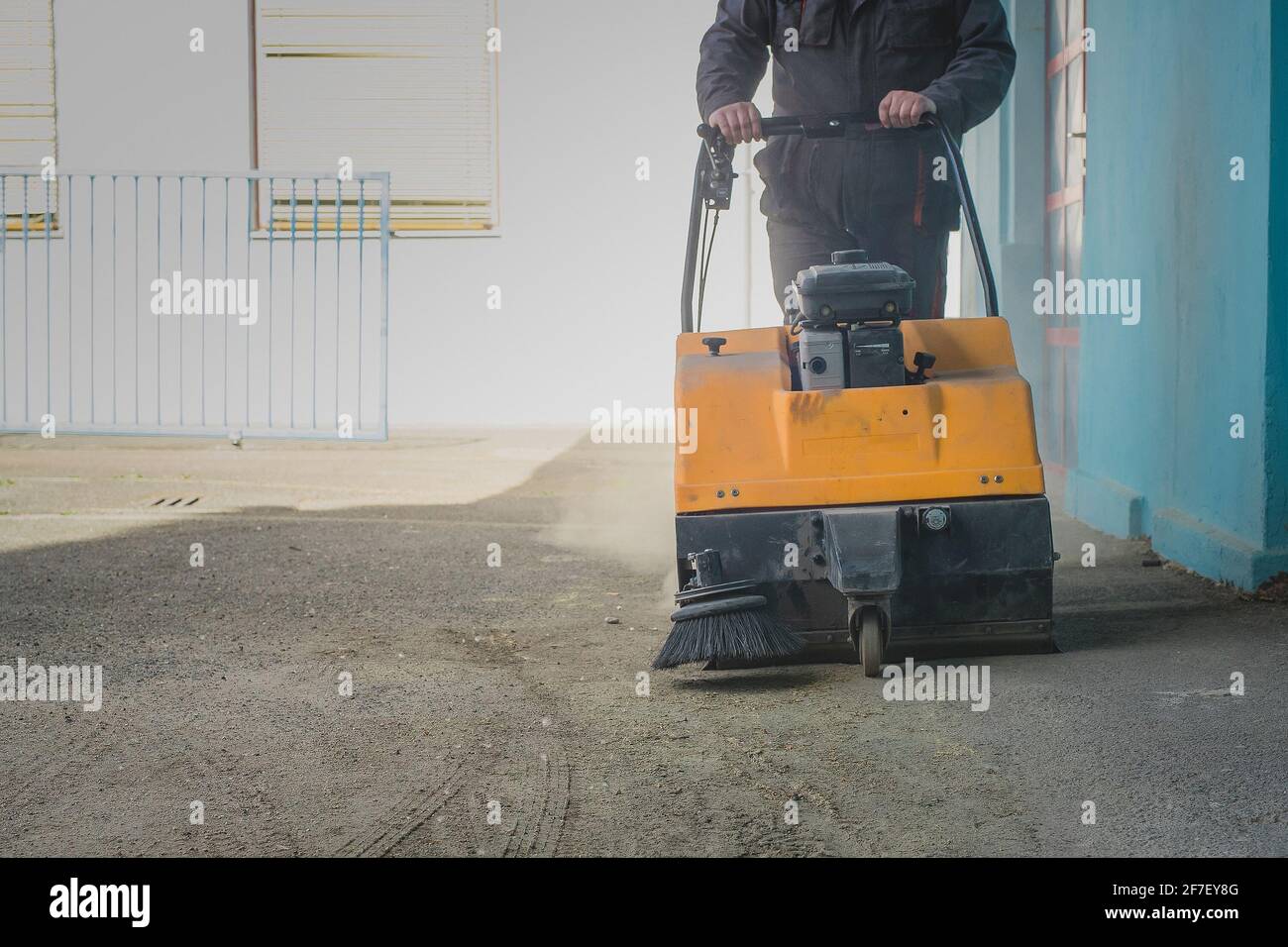 Una persona sta spingendo una spazzatrice industriale alimentata a benzina arancione su asfalto sporco nel tentativo di pulirlo da detriti e sabbia Foto Stock
