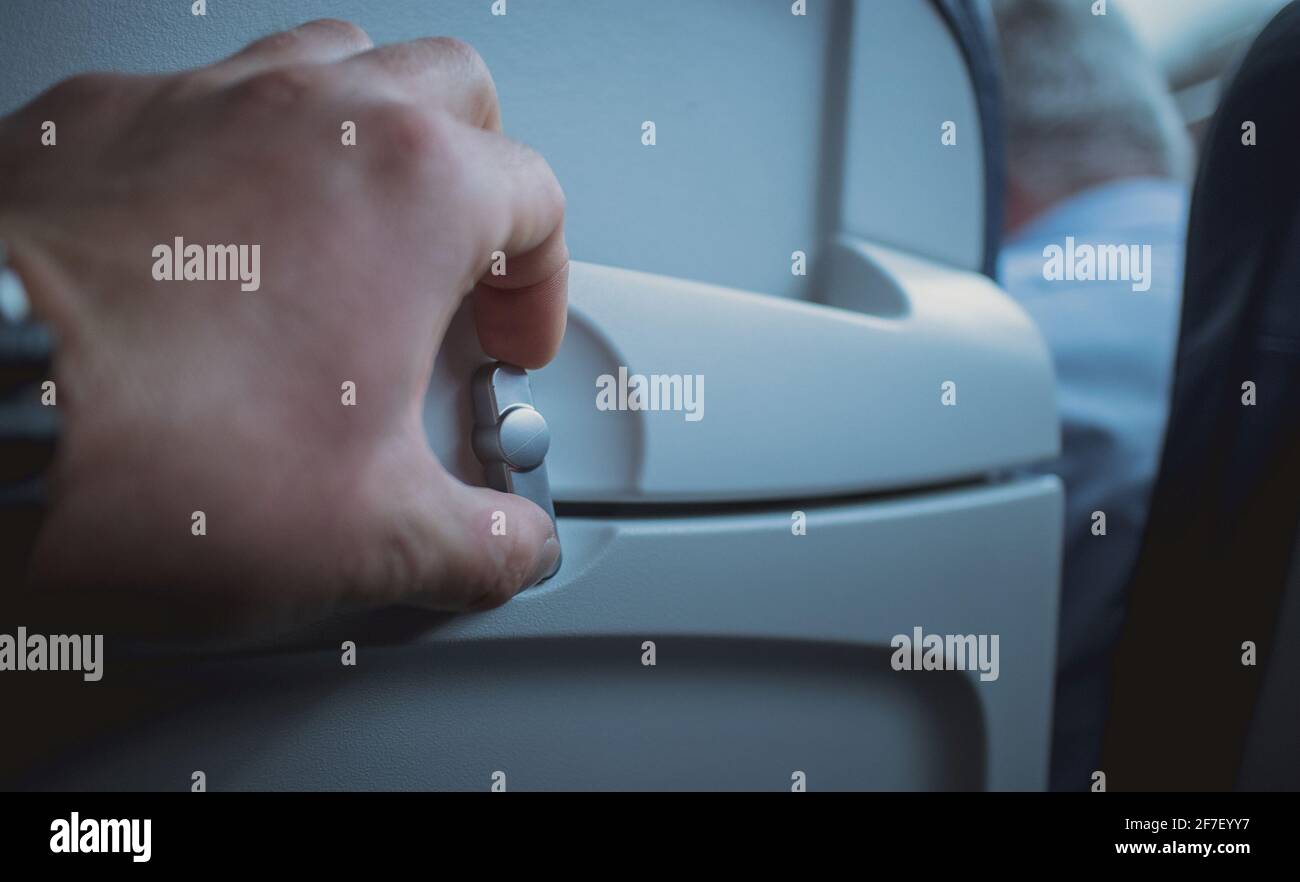 Manopola di apertura manuale per il rilascio del vassoio del tavolo sullo schienale di un sedile aereo. Vassoio visibile in posizione di stivaggio con una mano di una persona che ruota la manopola. Foto Stock