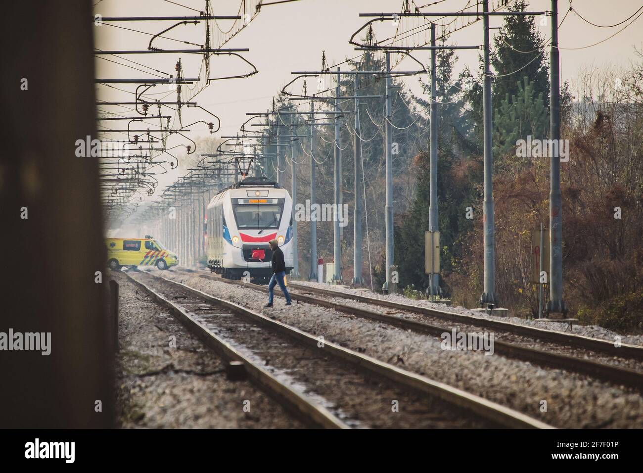 Uomo ignorante che cammina sulle piste del treno con il treno bianco per passeggeri in avvicinamento. Un'ambulanza sta attraversando i binari dietro il treno. Concetto di Foto Stock