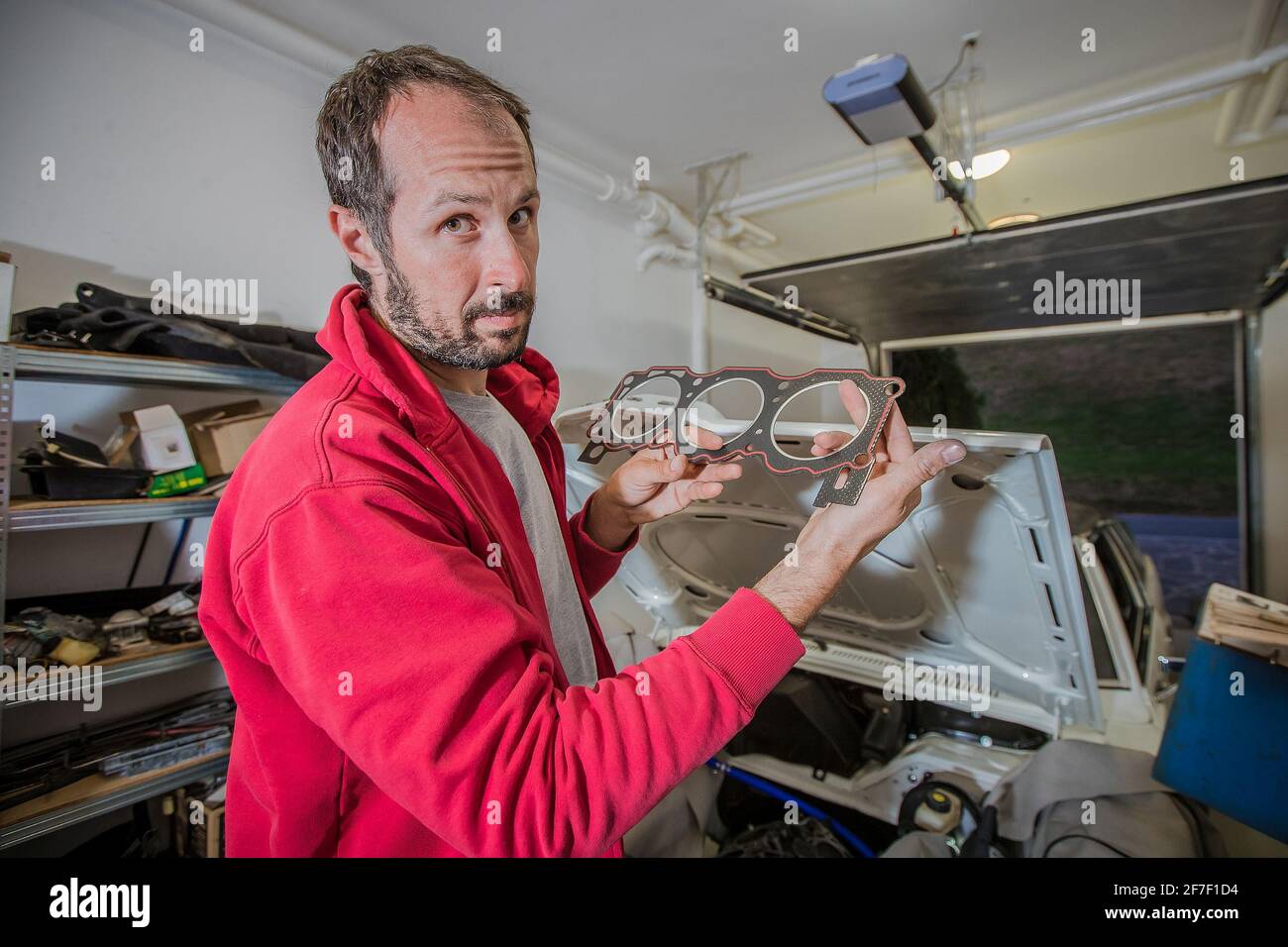 Un meccanico domestico che monta una testata V6 nel garage. Meccanico che tiene una testa a tre cilindri givata tra le braccia. Vettura con cofano aperto vista nella Foto Stock