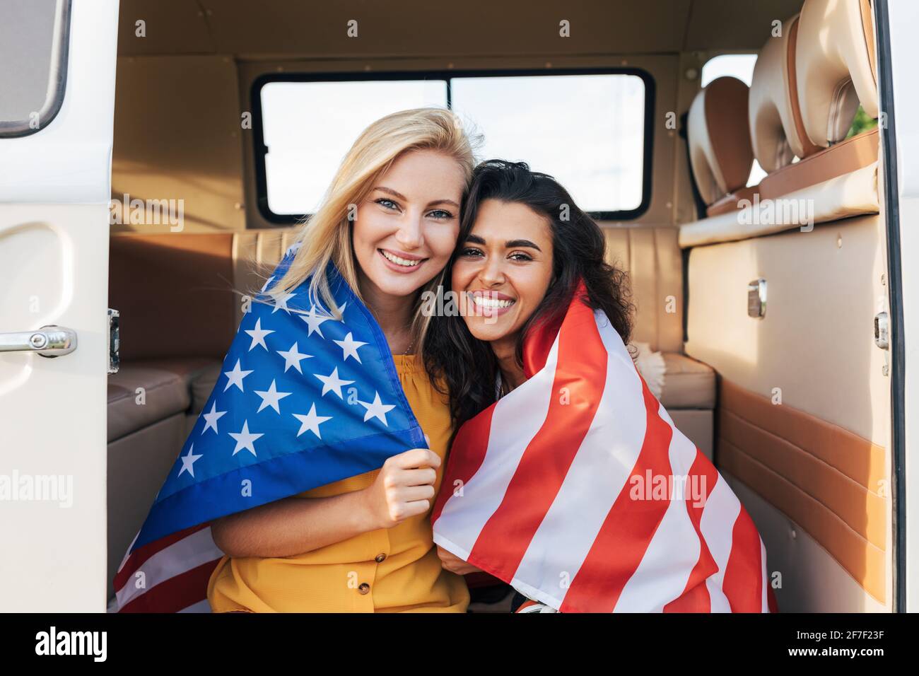 Due giovani amici femminili con bandiera americana seduti in un camper. Belle donne con bandiera degli Stati Uniti Foto Stock