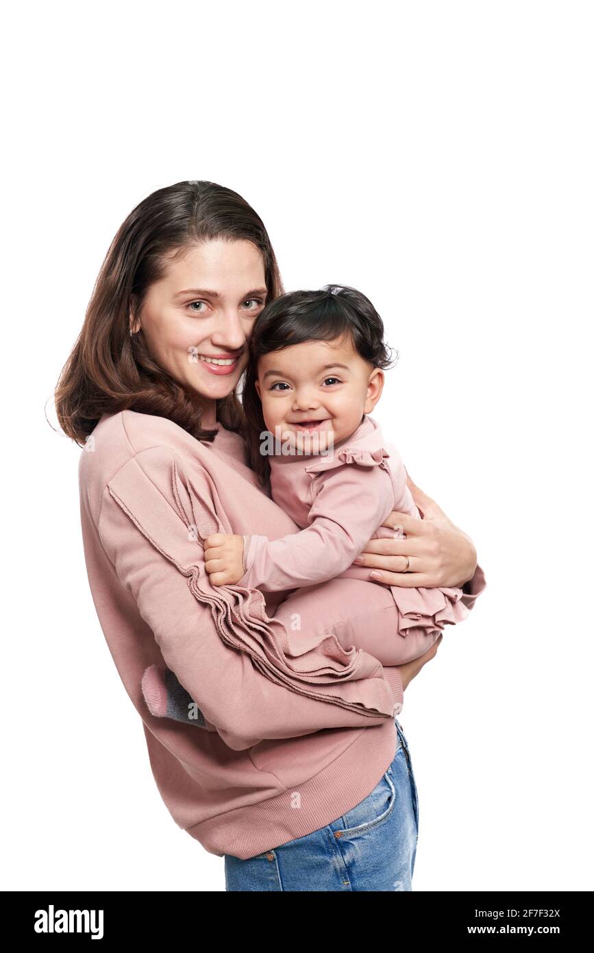 Ritratto di carina madre che tiene figlia isolato su sfondo bianco studio. Vista laterale di una giovane donna attraente che abbracciano un bambino dolce adorabile mentre si posa e si guarda la macchina fotografica. Foto Stock