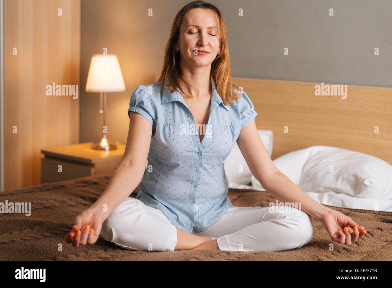 Ritratto di donna sorridente tranquilla che si rilassa sulla posizione di lotus seduta sul letto in camera da letto accogliente e guardando la macchina fotografica. Foto Stock