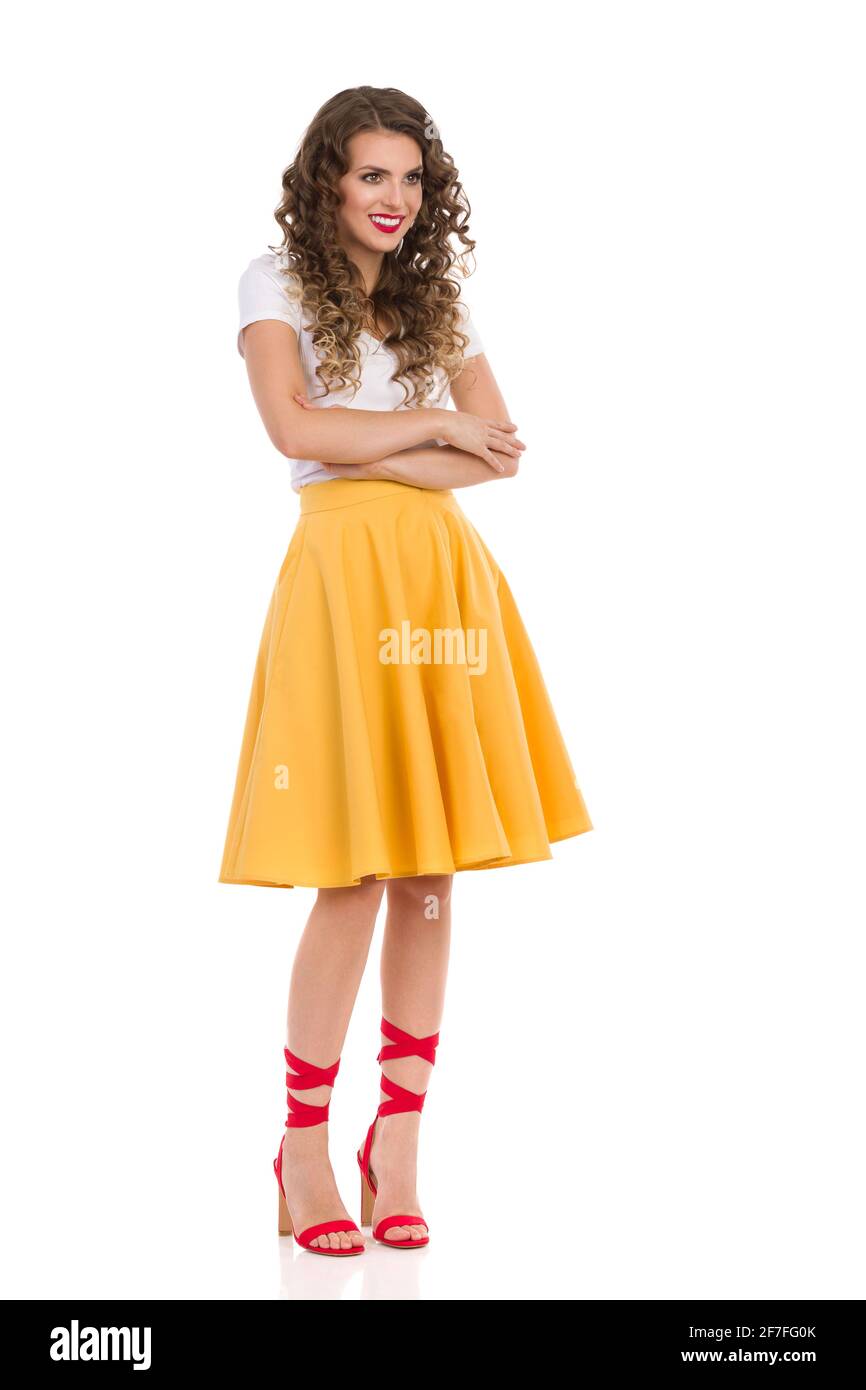 Bella giovane donna con gonna gialla, top bianco e tacchi rossi alti è in piedi con le braccia incrociate, guardando via e sorridendo. Foto in studio a lunghezza intera i Foto Stock