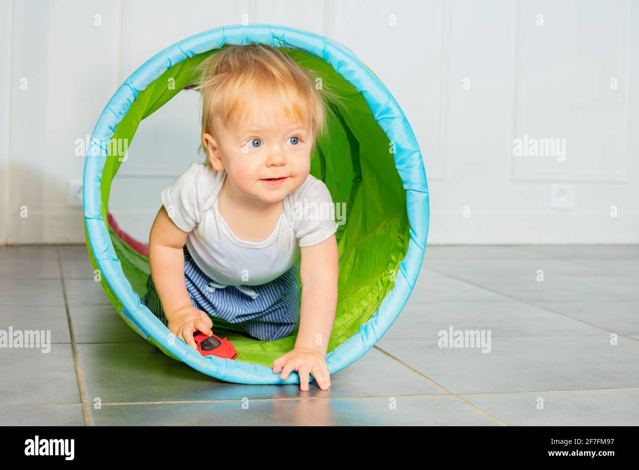 Il ragazzo biondo del toddler strisciano nel tubo con l'automobile giocattolo Foto Stock