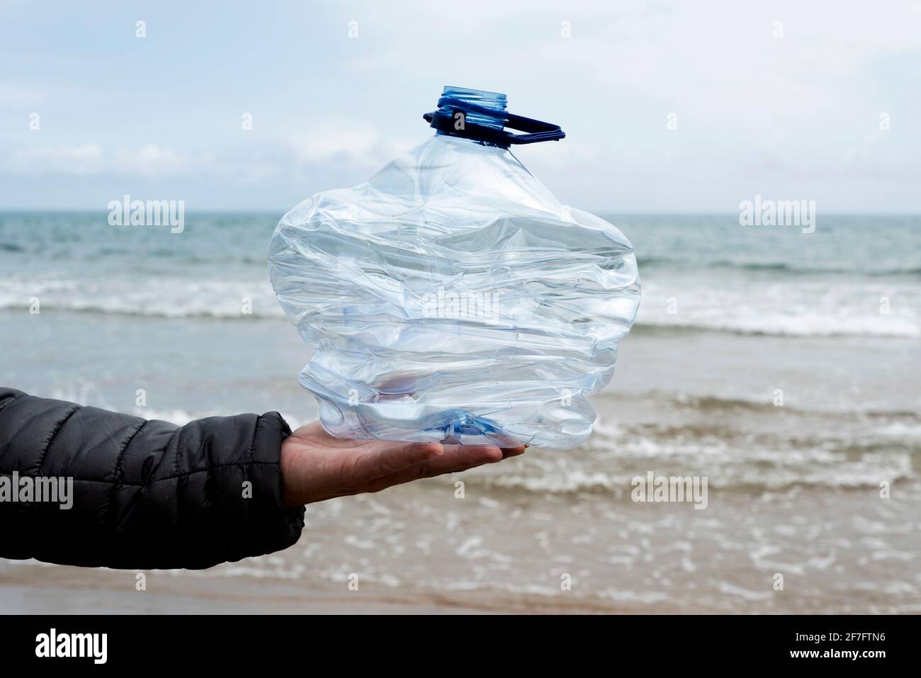 primo piano di un giovane uomo caucasico di fronte al ocean tenendo una bottiglia di plastica usata in mano di fresco raccolto dalla sabbia di una spiaggia Foto Stock