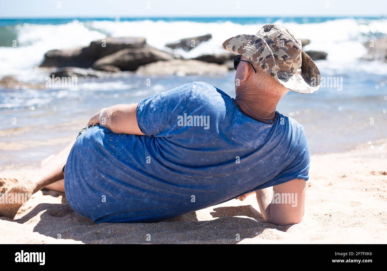 Un uomo europeo adulto si trova sulla spiaggia sulla sabbia e guarda le onde che si infrangono contro le rocce. Viaggi e turismo. Foto Stock