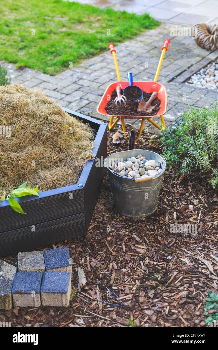 Giardinaggio - compostore, secchio con pietre e carriola con terreno. Letto con pacciame e prato verde. Foto Stock