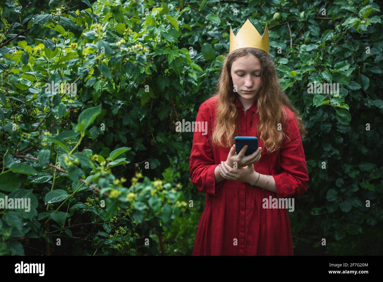 Presteen ragazza che indossa abito rosso e corona di carta in piedi giardino bagnato utilizzando il telefono Foto Stock