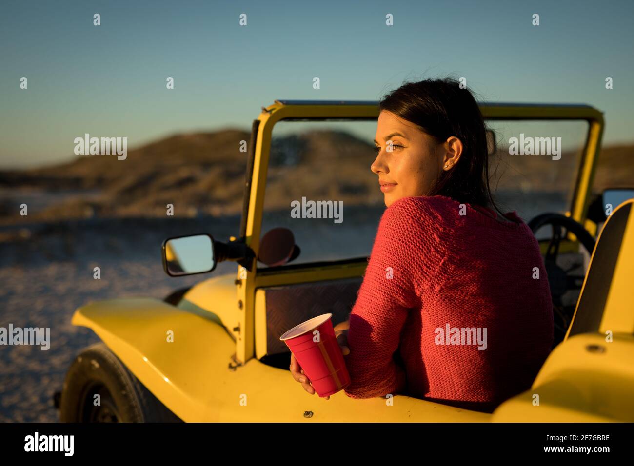 Donna caucasica felice seduta in buggy spiaggia vicino al mare tenendo la tazza rossa Foto Stock