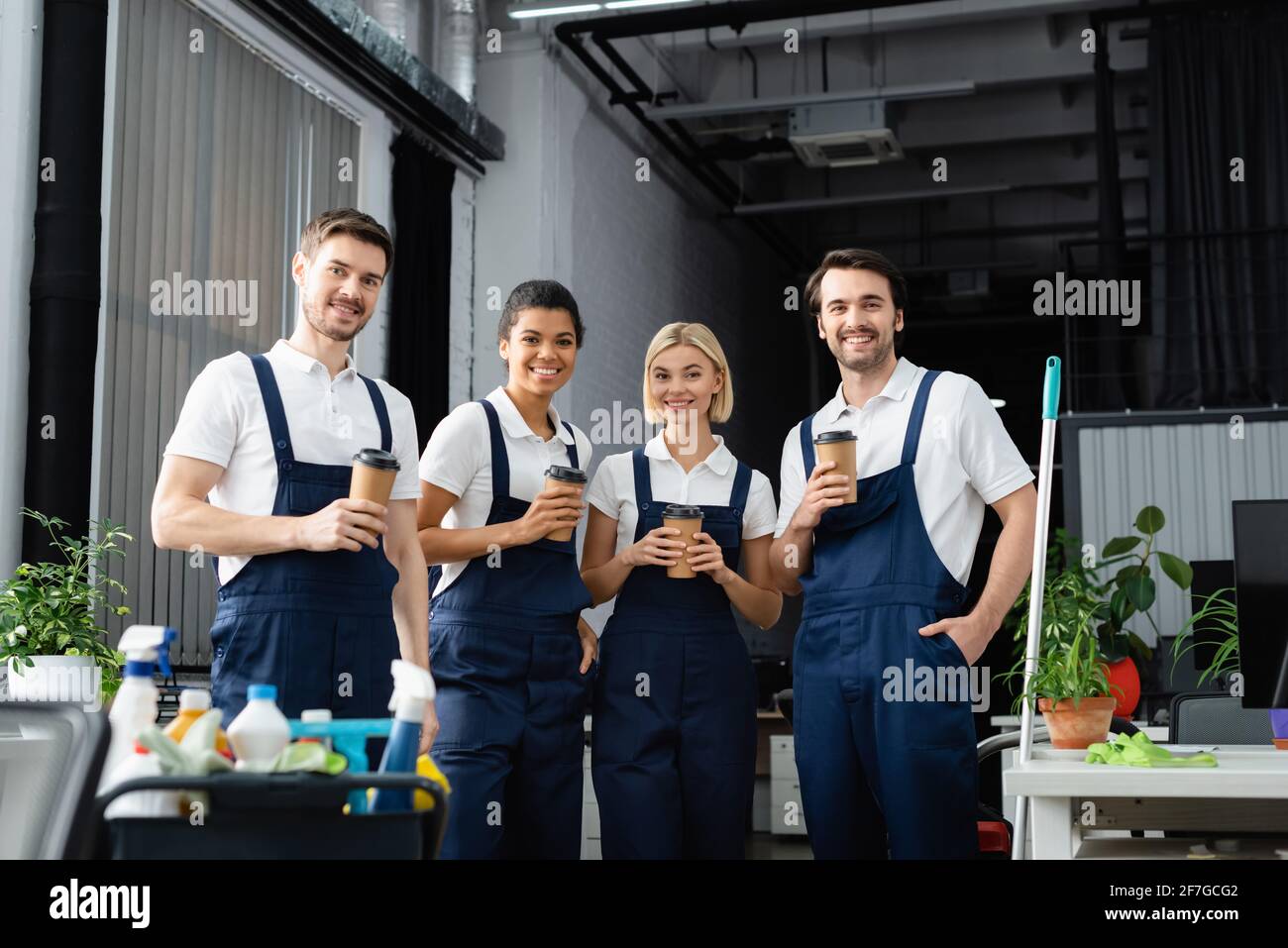 Detergenti per uso interrazziale con tazze di carta che guardano la fotocamera vicino ai detergenti in primo piano sfocato in ufficio Foto Stock