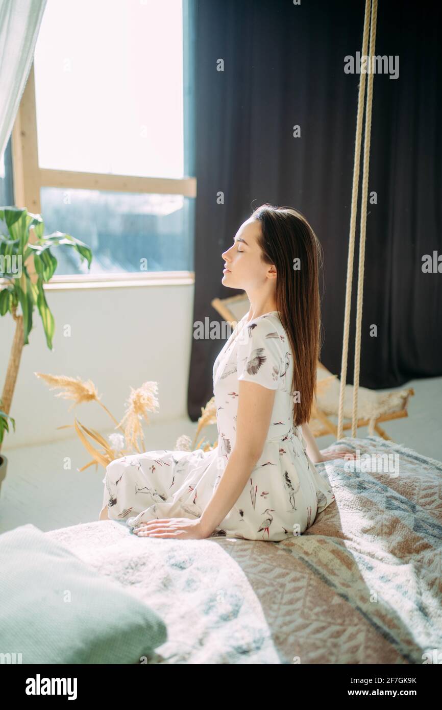 La giovane donna si siede sul letto in vestaglia e medita accanto alla finestra. Foto Stock