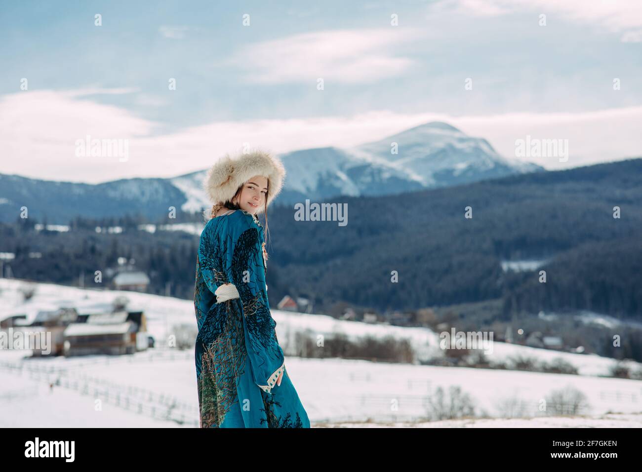La giovane donna cammina vestita di cappello di pelliccia e vestirsi contro il bellissimo paesaggio di montagne, cielo e villaggio dei Carpazi innevati. Foto Stock