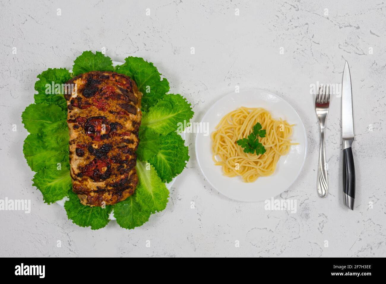 Un grosso pezzo di maiale cotto in forno, cotto in forno, su foglie di lattuga verde. Con la pasta su un piatto con una forchetta e un coltello. Foto Stock