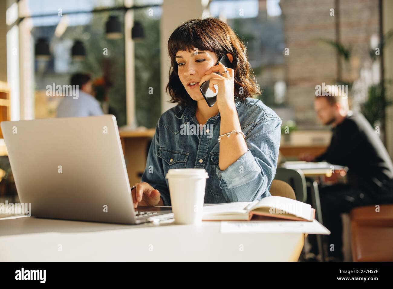 Giovane freelance seduto in uno spazio di coworking che parla sul telefono cellulare e utilizzando il computer portatile. Donna che usa il laptop e parla con il cliente sul telefono cellulare. Foto Stock