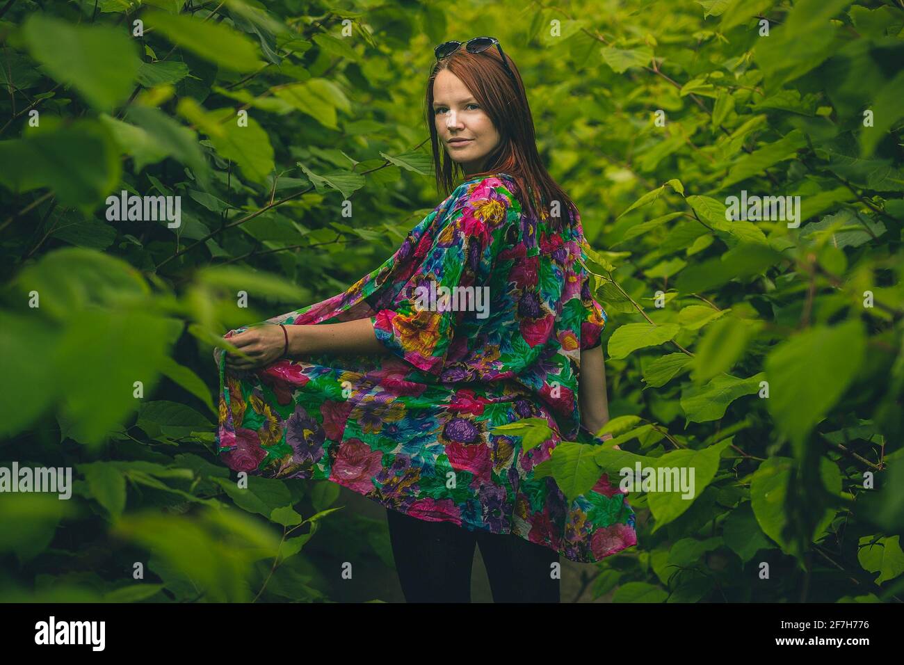 Donna in un abito colorato in posa in una giungla verde lussureggiante come foresta o cespuglio e girando la testa verso la fotocamera. Foglie che circondano una bella ragazza rossa exp Foto Stock