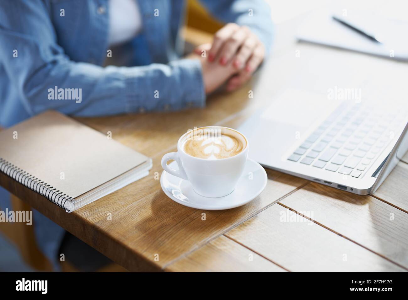 Primo piano di donna seduta al tavolo con una tazza di caffè gustoso, computer portatile moderno e notebook con penna. Concetto di preparazione al lavoro in caffetteria con computer portatile. Foto Stock