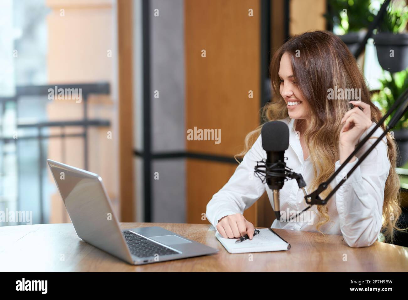 Vista laterale di una bella donna sorridente in camicia bianca che comunica con il microfono online dal computer portatile in caffetteria. Concetto di processo che parla con i seguaci online con buon umore. Foto Stock