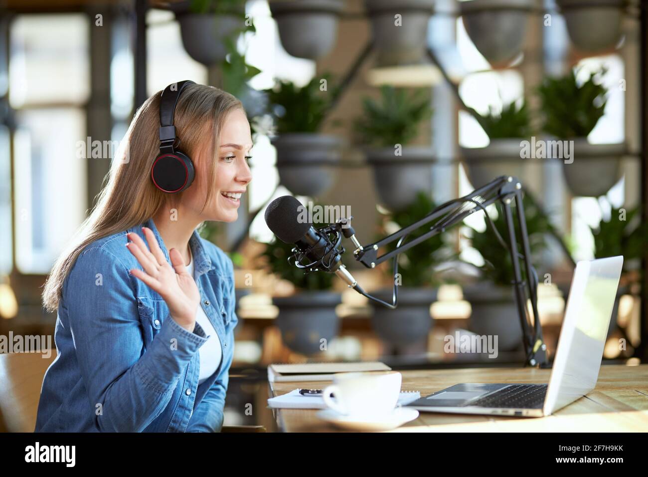 Vista laterale della giovane sorridente bella blogger donna che parla con i seguaci o gli amici online in cuffia sul computer portatile. Concetto di processo di comunicazione online con caffè gustoso. Foto Stock