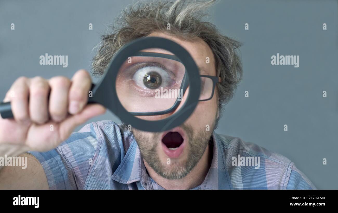 Un curioso e divertente uomo di mezza età sorpreso che sta guardando la fotocamera attraverso una lente di ingrandimento. Umoristico ritratto di un uomo pazzo senza capelli tagliati. Foto Stock