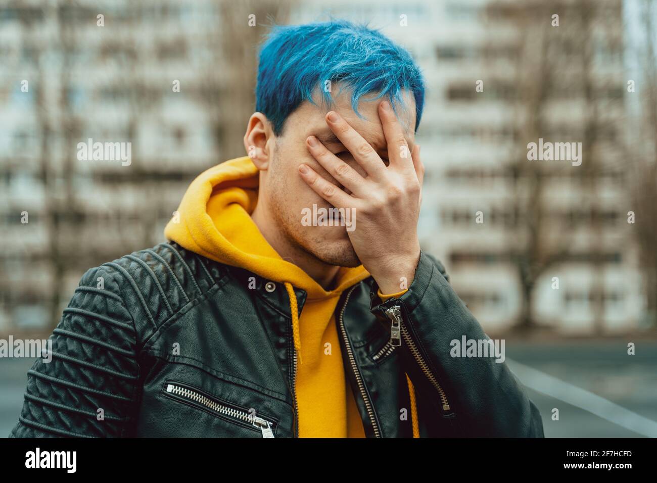 Ritratto di giovane uomo che chiude il volto con la mano sullo sfondo di un alto edificio. Bel ragazzo con i capelli blu che si posano sulla strada della città in primavera Foto Stock