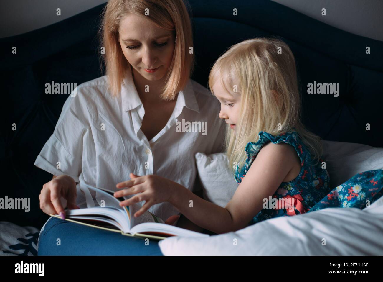 Felice famiglia di mamma bionda e figlia stanno leggendo un prenota a letto Foto Stock