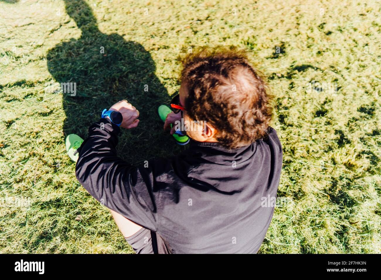 Corridore uomo di mezza età seduto sull'erba in un parco indossare gli occhiali guardando lo smartwatch al piano di allenamento Foto Stock