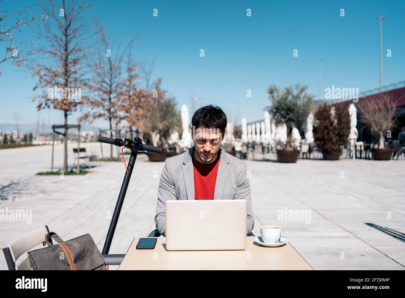 Giovane imprenditore che lavora sul computer all'aperto prendendo un caffè con uno scooter elettrico in piedi accanto a lui Foto Stock