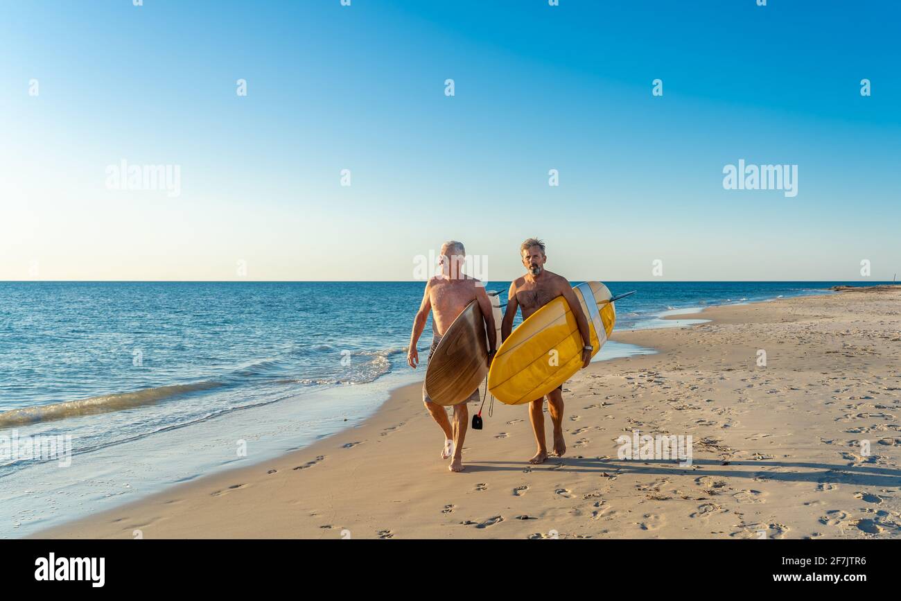 Due uomini maturi che camminano con tavole da surf sulla bellissima spiaggia godendo di paradiso e di vita di riposo. Amici adulti in forma attraente che si divertono Foto Stock