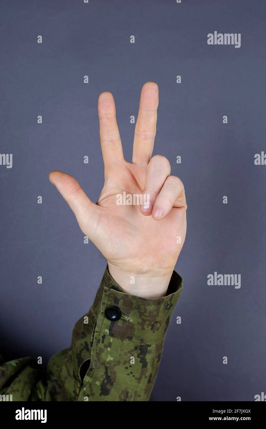 Mano in camuffamento mostra gesto con le dita su sfondo grigio. Tre dita sono sollevate. La designazione del numero tre nell'esercito Foto Stock