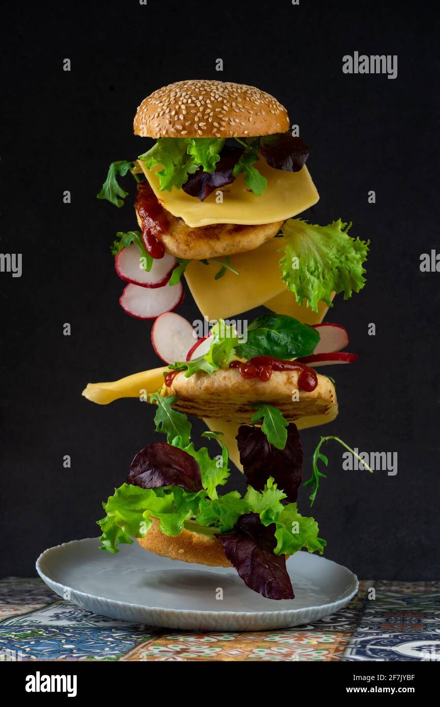 Levitating fette di hamburger. Cibo di volo. Hamburger Bun, cotoletta di pollo doppia con formaggio, lattuga, rucola e rafano. Carne, verdure e pane. Ingredienti su sfondo nero. Foto Stock