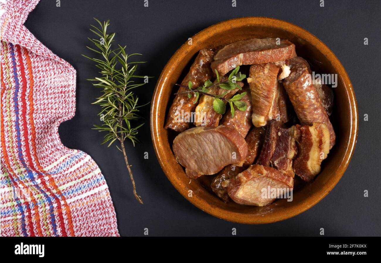Costolette di maiale conservate, lombo e salsiccia di maiale in una pentola di argilla con uno sprig di rosmarino su sfondo nero, tipica cucina spagnola Foto Stock