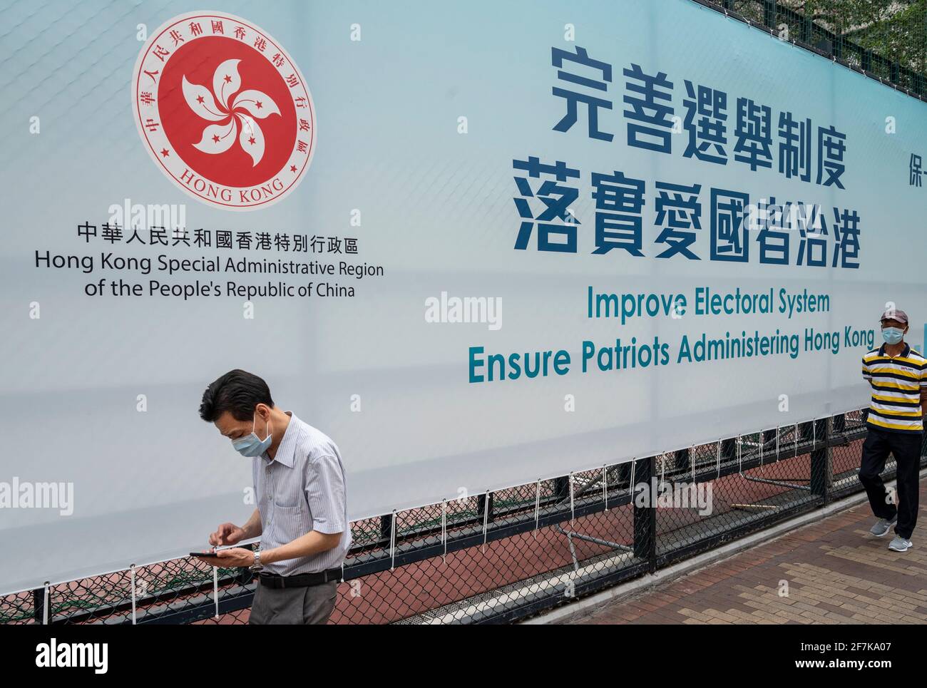 Le persone che indossano maschere facciali passano davanti a un banner governativo appeso su una recinzione che recita "migliorare il sistema elettorale garantire i patrioti che amministrano Hong Kong". Foto Stock