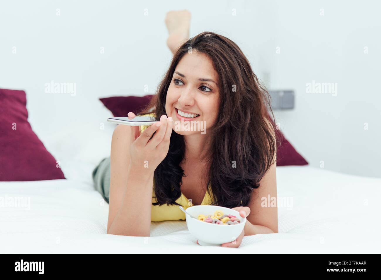 Donna sdraiata a letto con una ciotola di cereali durante l'invio di un messaggio vocale. Concetto di stile di vita. Foto Stock