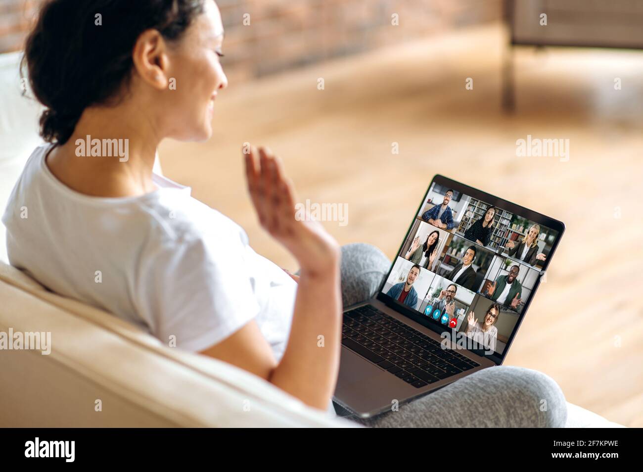 Lavoro remoto, comunicazione online. Una donna incinta usa un computer portatile, comunica con i colleghi tramite videochiamata mentre si siede a casa sul divano, saluta gli interlocutori con un gesto della mano, sorride Foto Stock